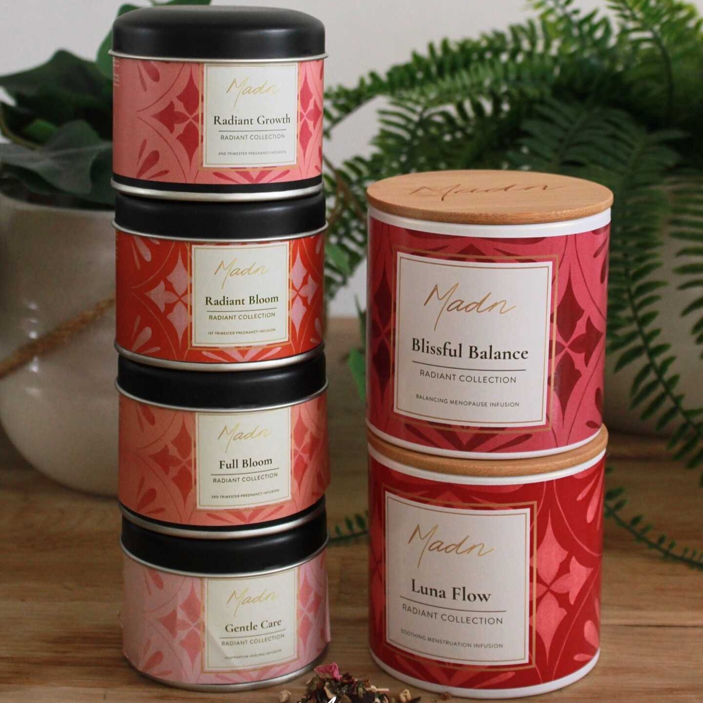 Stack of MADN Radiant Collection tea tins on a wooden table—Radiant Bloom, Radiant Growth, Full Bloom, Gentle Care, plus larger canisters of Blissful Balance and Luna Flow—with loose tea leaves in front and fern leaves in the background.